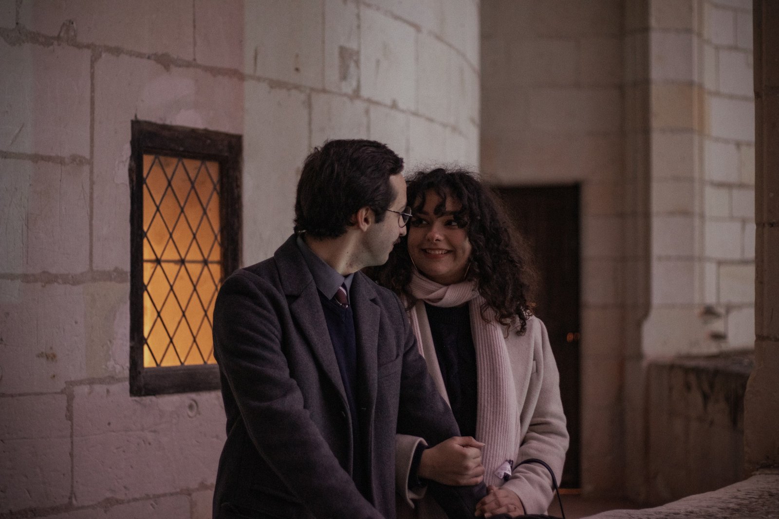 Couple standing close together in front of historic stone architecture with warm window light