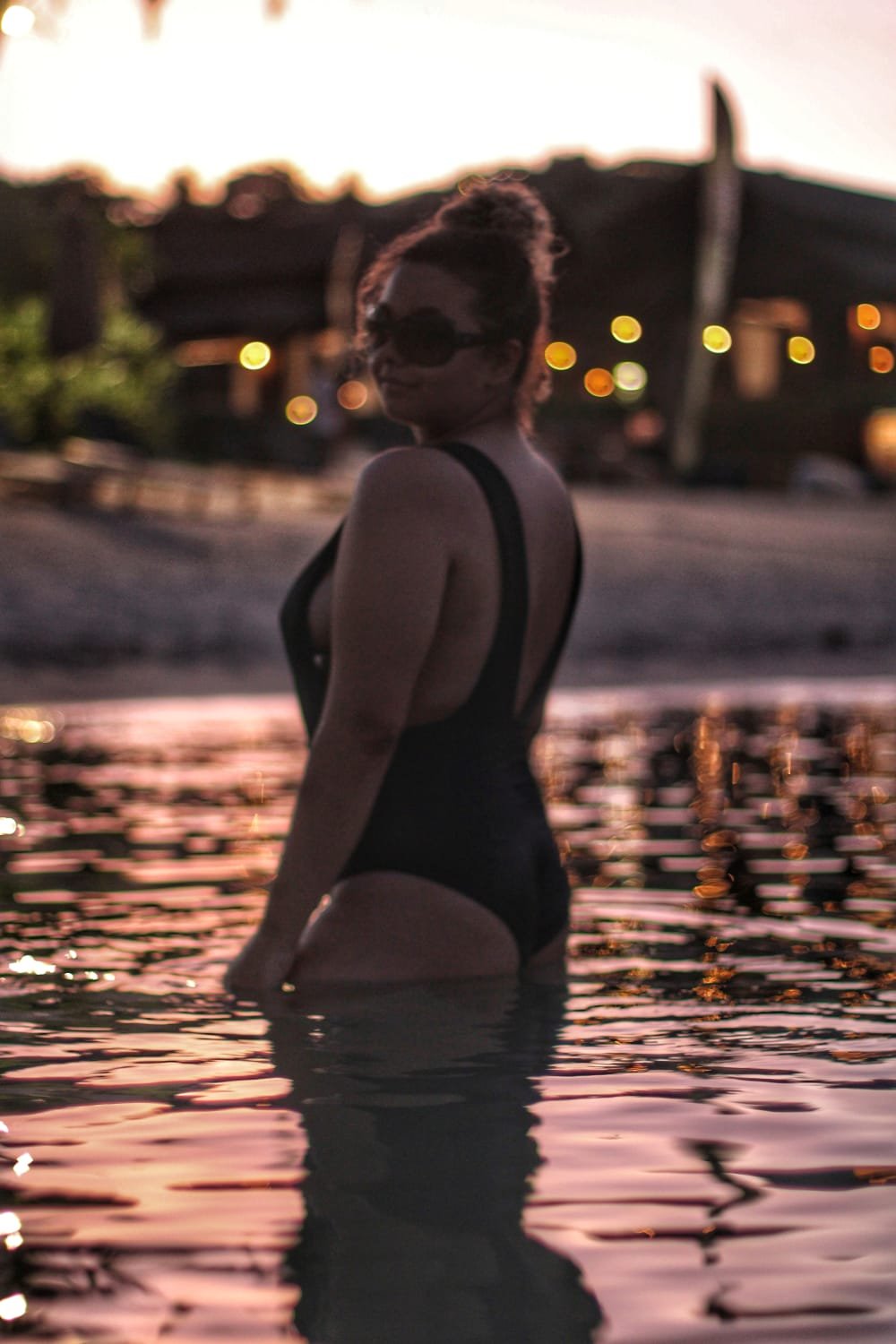 Woman standing in a swimming pool at sunset with warm lights in the distance